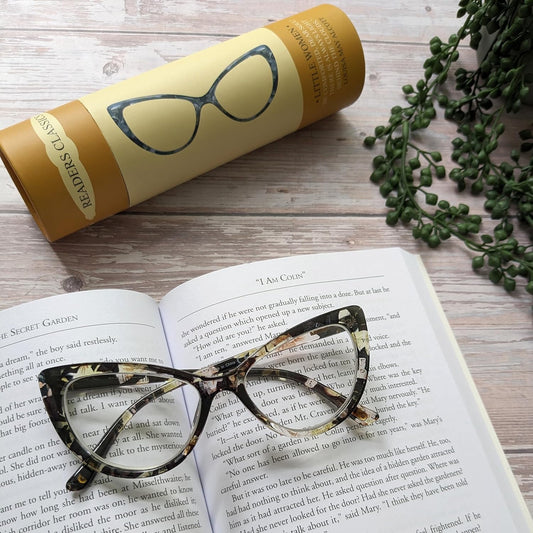 Cat-eye glasses on an open book with a yellow tube labeled 'Readers Classics' and greenery in the background.