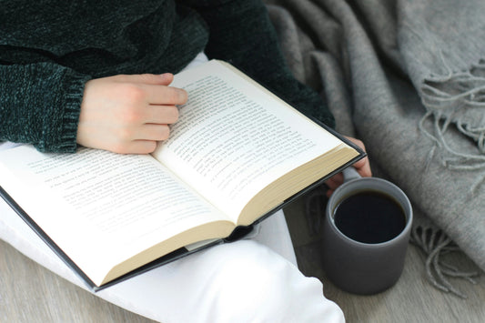 A person reading an open book with a dark mug beside them, wrapped in a cosy grey throw