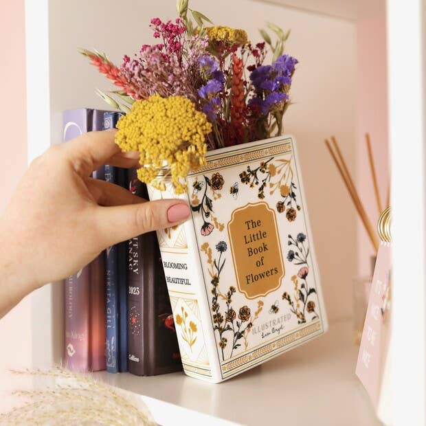 Hand holding a small jar of flowers with 'The Little Book of Flowers' book against a blurred bookshelf background.
