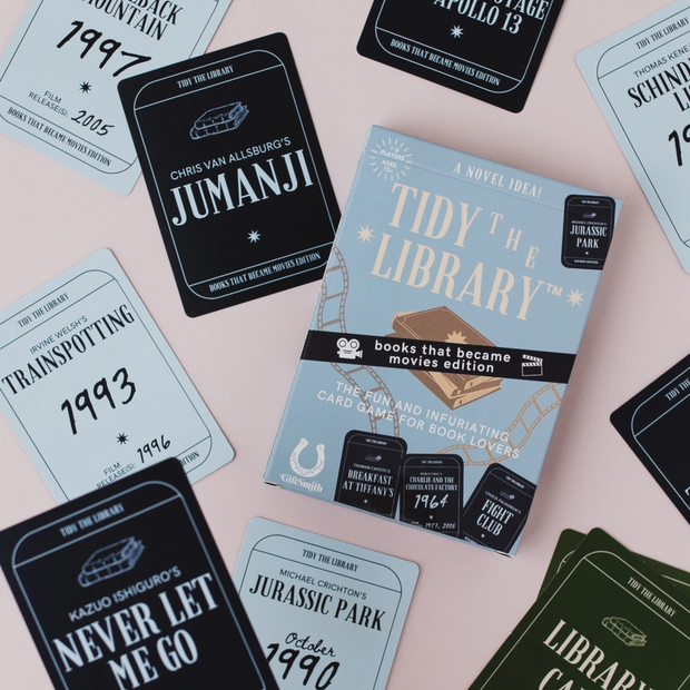 A boxed card game called "Tidy the Library" is arranged on a pale pink background with several sample card designs around it. The box is light blue with black and white typography and a small brown book illustration.