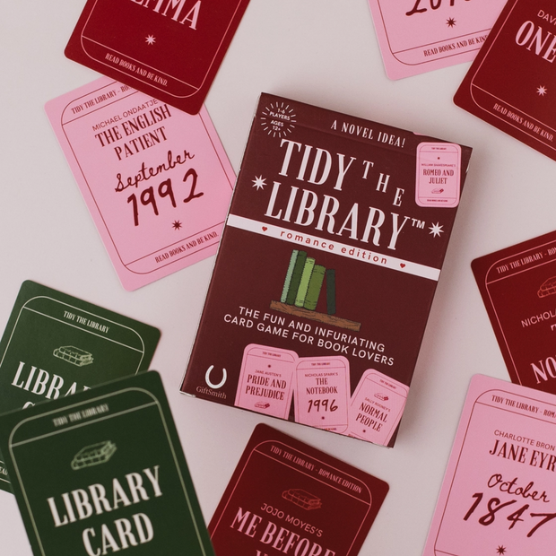 A boxed card game called "Tidy the Library" surrounded by matching red, pink, and green card sleeves on a light background. The box is dark brown with white and pink text, and the surrounding cards show different romance-themed library card designs.