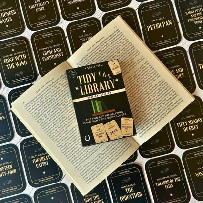 The image shows the card game 'Tidy the Library' laid out on an open book. The game includes a rulebook, library cards, and book cards with various book titles.